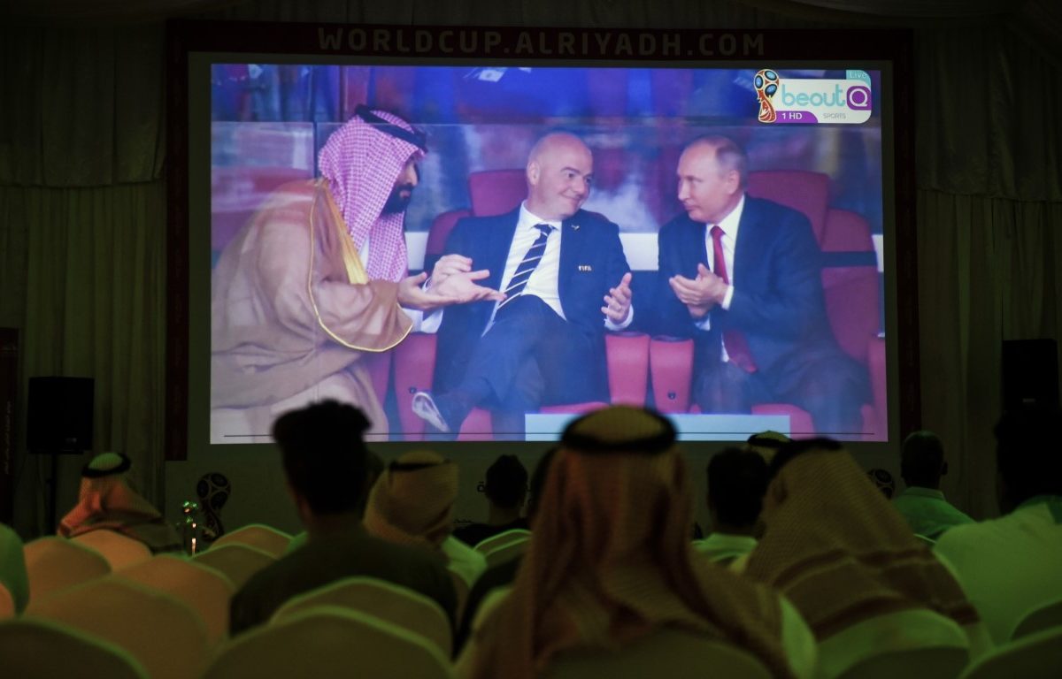 A picture taken on June 14 2018 in a Saudi football fan tent in the capital Riyadh shows a projector showing the Russia 2018 World Cup Group A football match between Russia and Saudi Arabia with a clip of Saudi Crown Prince Mohammed bin Salman L gesturing as he sits next to FIFA President Gianni Infantino C and Russian President Vladimir P